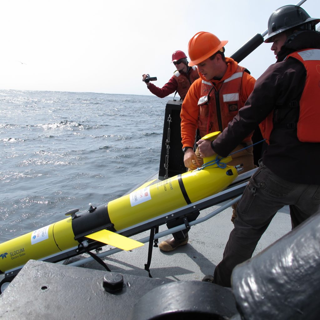 Two scientists in work vests and safety gear deploy a yellow glider off the deck of a vessel while being filmed by another person.