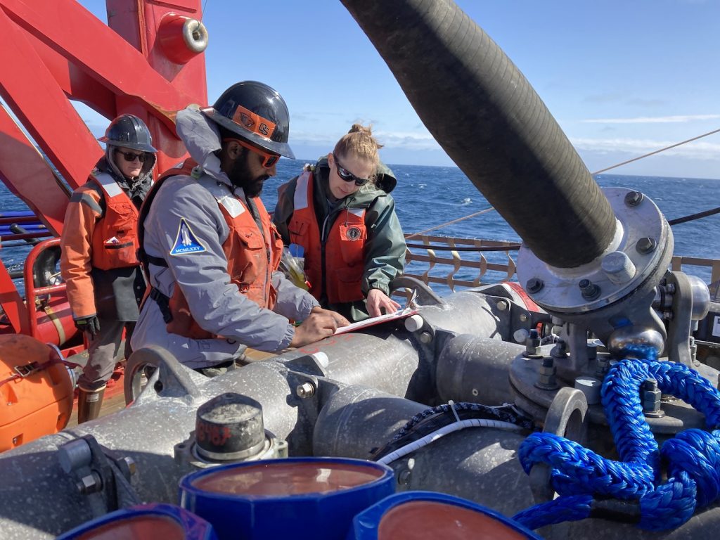 Three scientists in work vests and safety gear stand over a mooring sensor package on deck of a ship.