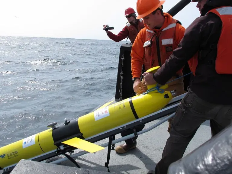 Two scientists in work vests and safety gear deploy a yellow glider off the deck of a vessel while being filmed by another person.