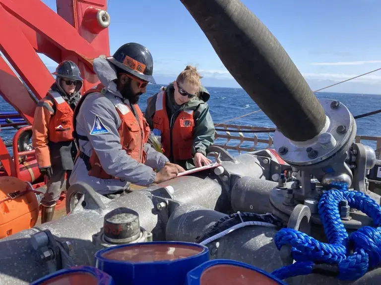Three scientists in work vests and safety gear stand over a mooring sensor package on deck of a ship.